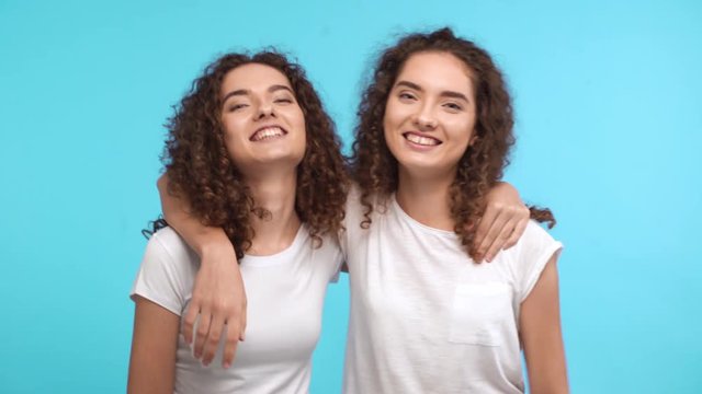 Two charming Caucasian curly female twins in white t-shirts embracing each other on shoulders and showing ok with thumbs on blue background