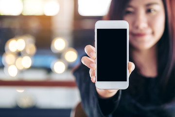 Mockup image of a beautiful woman holding and showing white mobile phone with blank black screen and smiley face in vintage cafe