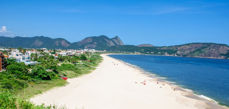 View of the beautiful Camboinhas Beach with houses, green trees, white sand, blue ocean and mountains on the background, Niteroi, Rio de Janeiro, Brazil