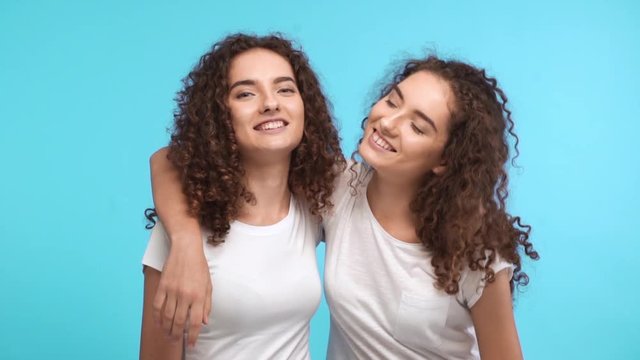 Two charming Caucasian curly female twins in white t-shirts embracing each other on shoulders smiling and showing ok with thumbs on blue background in slowmotion