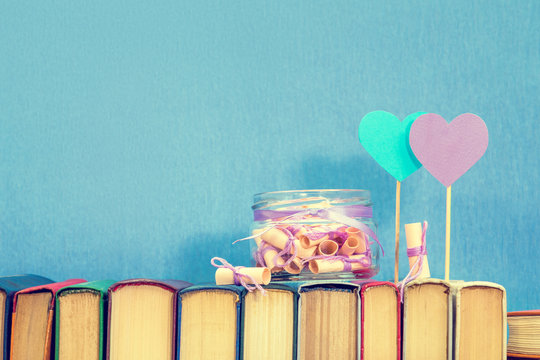 Valentines day concept. Dreams written on a pink rolled paper in a glass jar, blue paper heart, violet paper heart on books over blue background. Toned.
