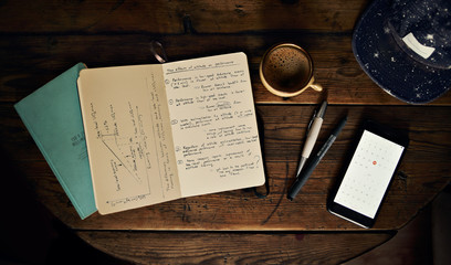 Notebooks, pens, smartphone, cup of coffee and a hat shot from above on a vintage brown table, top...