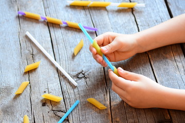 Development of fine motor skills for kids. Small kid holds a straw and a raw pasta in his hands. Creative kids activity. Easy exercise to improve hand and eye coordination. Wooden background