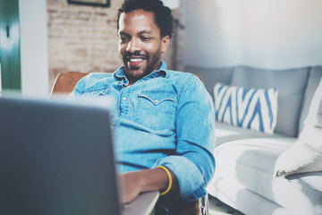 Smiling young African man making video conversation with friends while sitting on sofa at his sunny home.Concept of happy business people.Blurred background, film effect.