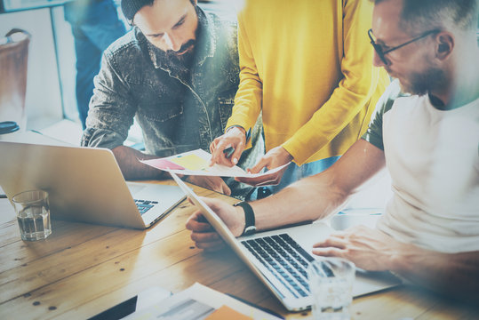 Coworking process in a sunny office.Two bearded mans working on computer at the wood table.Woman wearing yellow pullover and showing document in her hand.Horizontal,blurred background