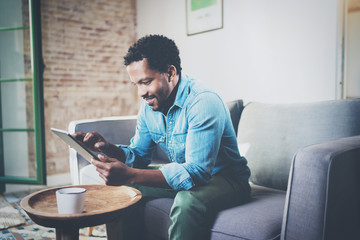 Attractive bearded African businessman using tablet while sitting on sofa at his modern home.Concept of young people working mobile devices.Blurred background. © SFIO CRACHO