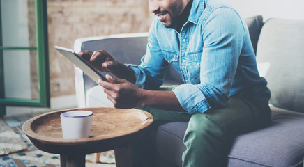 Attractive bearded African businessman using tablet while sitting on sofa at his modern home.Concept of young people working mobile devices.Blurred background.Crop. © SFIO CRACHO