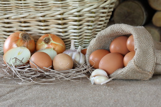 Куриные яйца, лук и чеснок лежат столе на фоне плетеной корзины и дров.