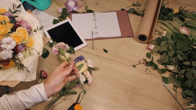 Male florist making bouquet at flower shop on wooden table, top view of hands with flowers, tablet, roses and decorating paper.