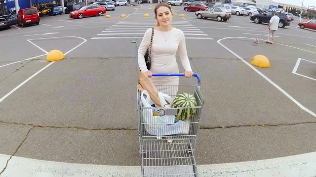 Beautiful young woman walking on parking lot at supermarket with full shopping cart