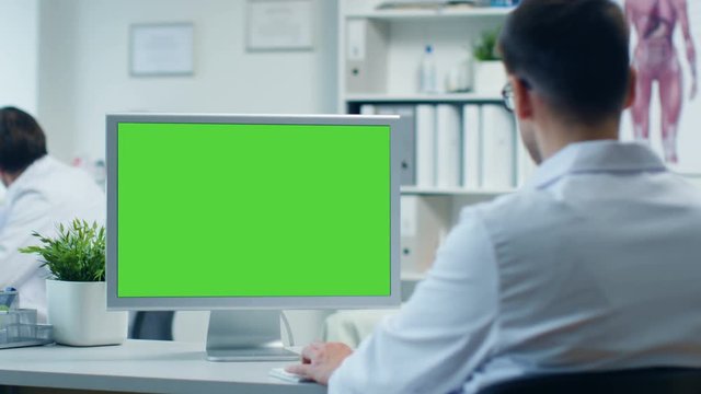 Doctor's Office. Male Doctor Works at His Personal Computer with Green Screen, His Assistant Comes in and Takes His Place at the Table. Office is Modern. Shot on RED Cinema Camera 4K (UHD).