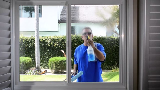 A man washes a window on his home using a spray bottle of window cleaner and a two sided squeegee to bring the glass to a sparkling clean finish. .
