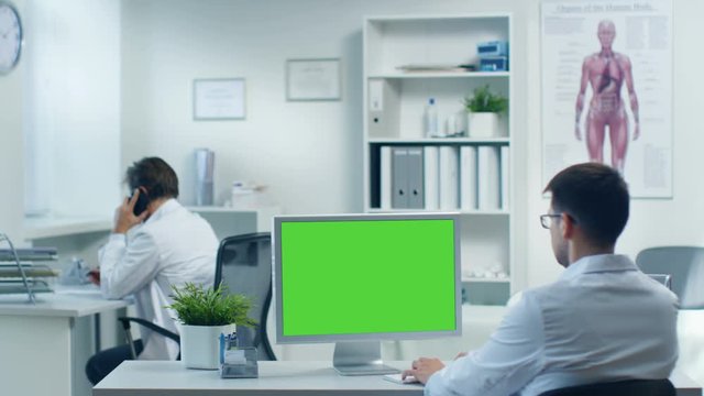 Male Doctor Working at His Personal Computer with Green Screen, His Assistants Consults Patients on the Phone. Shot in a Modern Medical Office. Shot on RED Cinema Camera 4K (UHD).