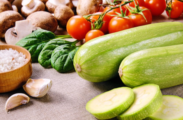 Two whole zucchini and a few sliced pieces, cherry tomatoes, mushrooms, salt in wooden salt shaker, cloves of garlic and spinach leaves on a linen cloth.