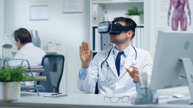 Male Doctor Conducting Experimental Medical Procedure Wearing Virtual Reality Headset. His Assistant Closely Monitors Activity from His Desk. Office is Modern. Shot on RED Cinema Camera