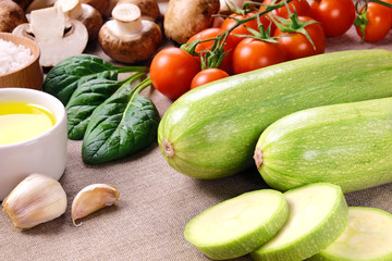 Two whole courgettes and a few sliced pieces, cherry tomatoes, mushrooms, spinach leaves, oil, salt and cloves of garlic on a linen cloth.