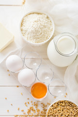 Ingredients for baking: flour, milk, wheat grain, butter and eggs on white wooden background. Selective focus
