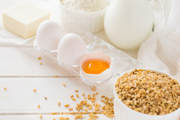 Ingredients for baking: flour, milk, wheat grain, butter and eggs on white wooden background. Selective focus
