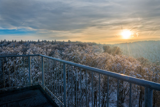 Beautiful sunset viewed over snow covered trees that extend to the horizon. The sun in its orange color is shining trough the cloud cover hitting the trees and railing in the foreground.