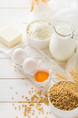 Ingredients for baking: flour, milk, wheat grain, butter and eggs on white wooden background. Selective focus
