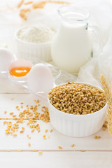 Ingredients for baking: flour, milk, wheat grain, butter and eggs on white wooden background. Selective focus
