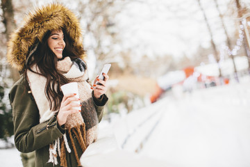 Young woman using a phone and drinking hot tea in the winter par