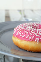 Light background with detail of sweet pink icing donut with sprinkles on gray plate. Selective focus