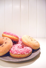 A ring donuts with pastel pink frosting and sprinkles on gray plate. White painted wooden planks on background. Selective focus. Place for your text