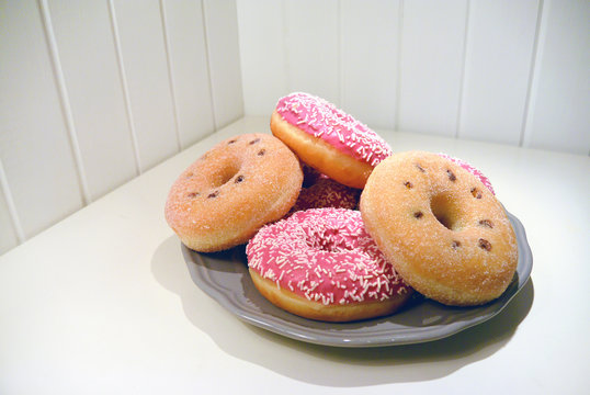 A ring donuts with pastel pink frosting and sprinkles on gray plate. White painted wooden planks on background.