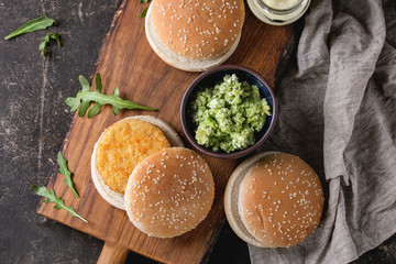 Ingredients for making vegan burger. Veggie cheese and onion cutlets, yogurt sauce, hamburger buns, herbs and avocado salad, served on wooden board with textile on dark background. Top view