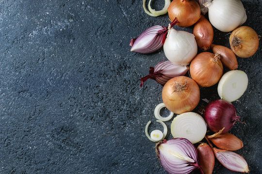 Variety of whole and sliced red, white, yellow and shallot onions over dark stone texture background. Top view, space for text