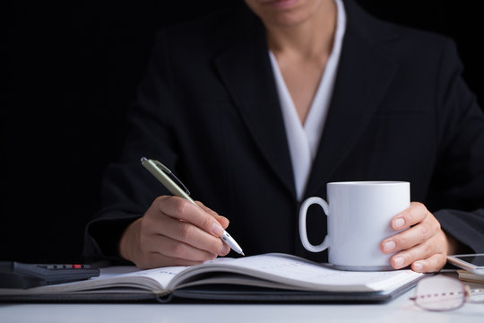 A business woman in black suit holding a white coffee cup in her hand and writing in the other hand on a working desk