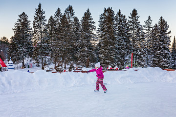 Little girl in warm clothes on an ice skating rink outdoors. Winter activity.