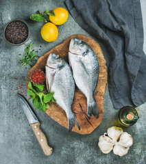 Fresh uncooked sea bream or dorado fish with lemon, herbs, olive oil, garlic and spices in bowls on rustic wooden board over grey concrete background, top view. Healthy, dieting, clean eating concept
