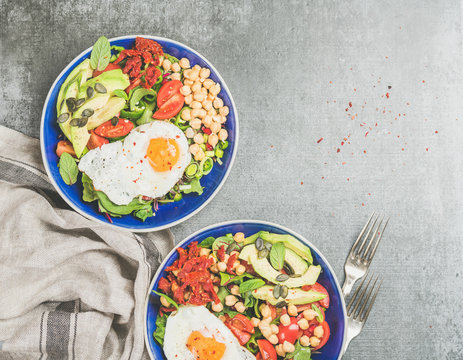 Healthy breakfast with fried egg, chickpea sprouts, seeds, vegetables and greens on bowls over grey concrete background, top view, copy space. Clean eating, healthy lifestyle, vegetarian food concept