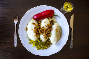 Potato dumplings - a traditional regional dish. Polish and Lithuanian cuisine. With pickles , fried bacon and paprika . White plate, knife, fork and glass of vodka on a dark background..