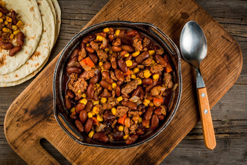 Traditional Mexican and Chilean dish of chilli con carne. In the rustic pan, next to tortillas and fresh vegetables on a wooden table, copy space