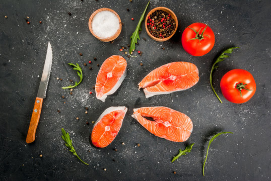 Raw uncooked salmon steaks on a dark gray stone kitchen table. With salt, spices, seasonings, herbs and vegetables for cooking. Top view, copy space