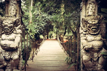 Fototapeta na wymiar Old wooden hanging bridge leading to tropical forest with terrible statues of Asian Buddhism demons on both sides in vintage style 
