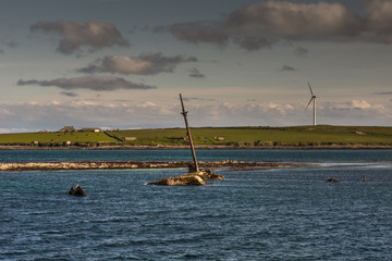 Orkneys, Scotland - June 5, 2012: The mast and other rusty parts of WW II warship wreck peep out of the sea near Lamb Holm Island. Wind turbine and farms. Small green band of land splits image in two