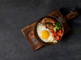breakfast in a frying pan, fried eggs, sausages and tomatoes on a dark background