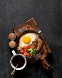 breakfast in a frying pan, fried eggs, sausages and tomatoes on a dark background
