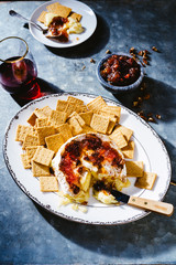 Brie cheese and cranberry pepper jelly party appetizer with crackers and wine on a zinc surface dark and moody lighting.