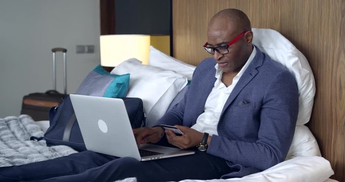 Online Shopping in the Hotel Room/Afro-american businessman is sitting in hotel bed, buying online. He’s happy to have this service, it’s very convenient. His suitcase is standing near