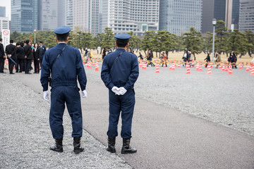 Here in the image two on duty Japanese police officers standing and watching people to maintain law and order in the area. And few people can be seen standing in a queue in the picture.