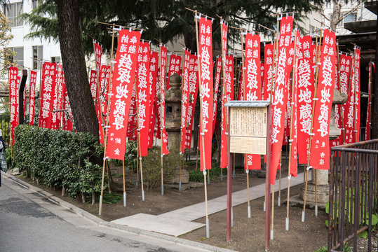 Lanterns with Japanese text written on it are seen on the road. On the background, lush green trees and the blue sky is seen on a sunny day. 
