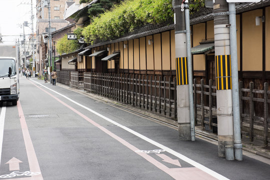 In this image we can see partially see a truck parked by the side of the road. Few green bushes can be seen on the roof of the buildings.