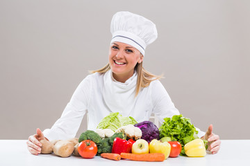 Cheerful female chef is sitting at the table with bunch of vegetable.

