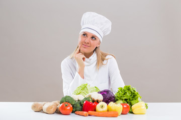 Cheerful female chef is sitting at the table with bunch of vegetable and thinking what to cook.
