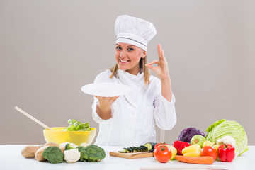 Portrait of cheerful female chef sitting at the table ,showing ok sign and empty plate while making healthy meal
